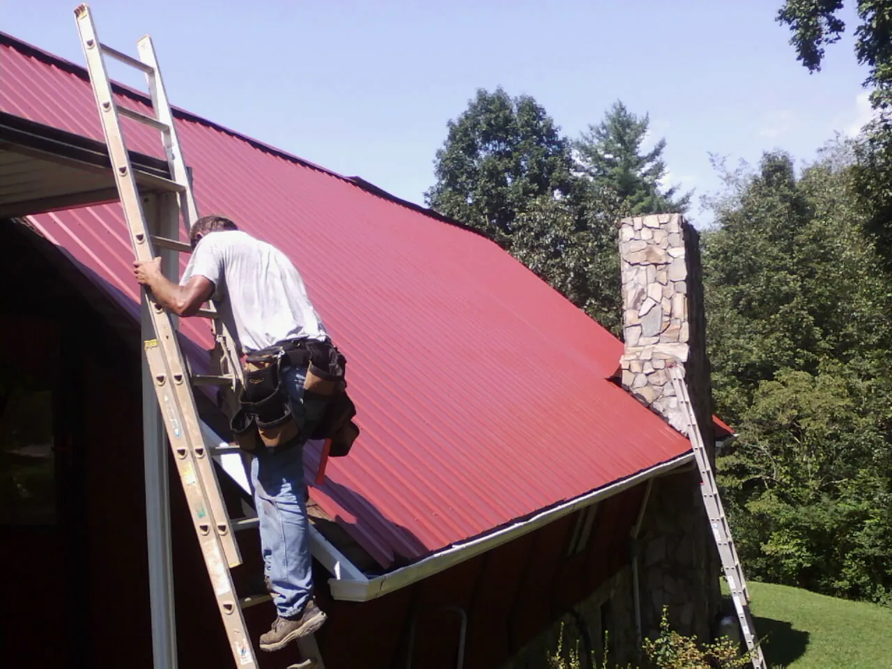 Roofer on a ladder beside a red metal roof and stone chimney — Soffit & Fascia Repair by Roofing Brothers in Fallon